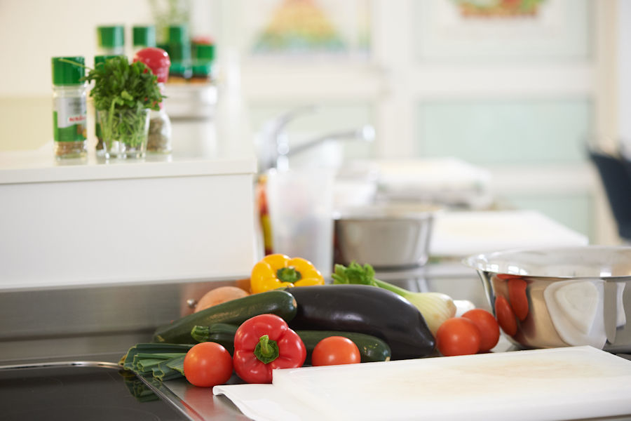 Buntes Gemüse auf einem Schneidebrett in der Lehrküche mit Gewürzen im Hintergrund Buntes Gemüse auf einem Schneidebrett in der Lehrküche mit Gewürzen im Hintergrund